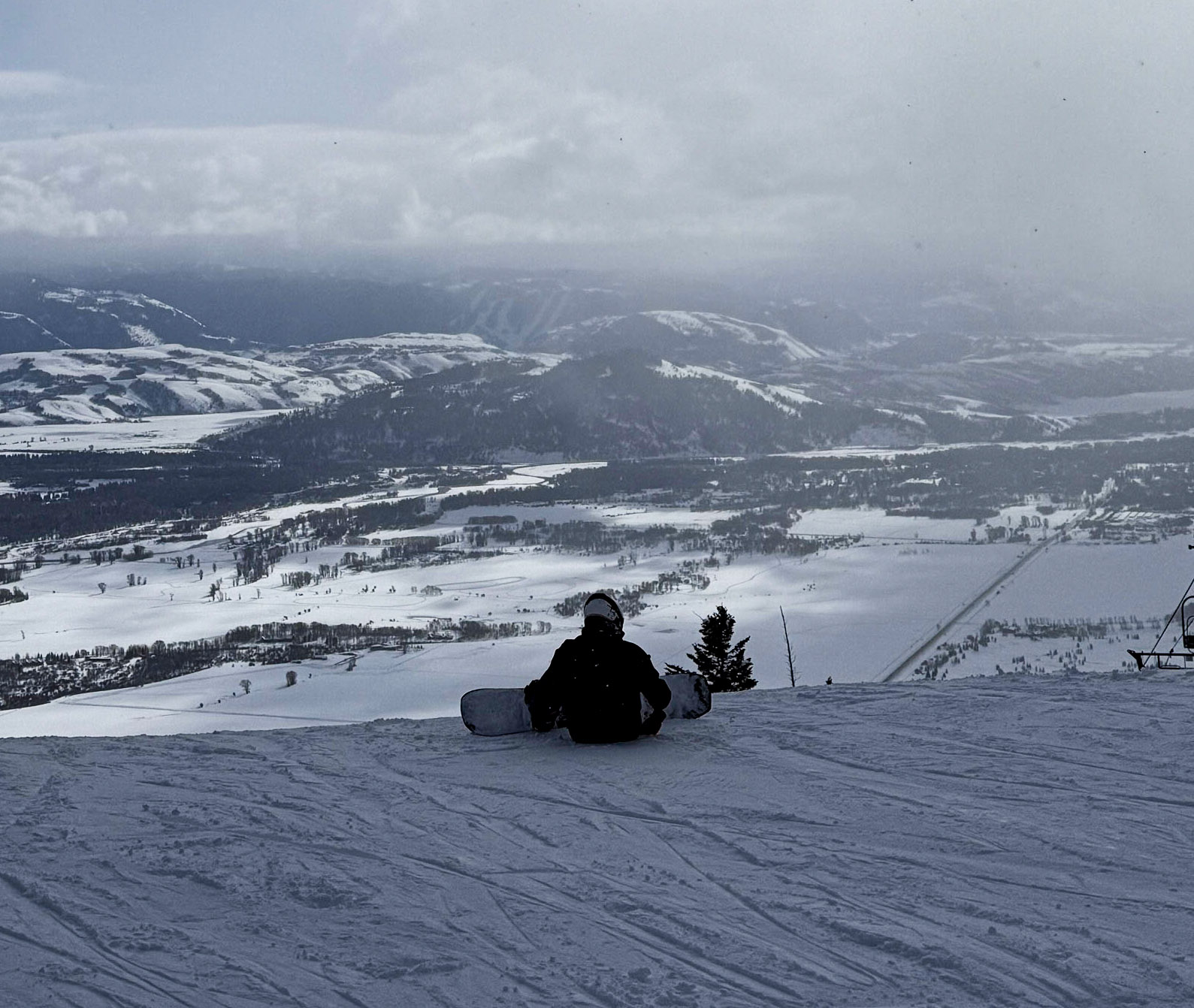 snowboarder sits at top of slope, with resort and horizon in the distant. Picture taken from behind the snowboarder